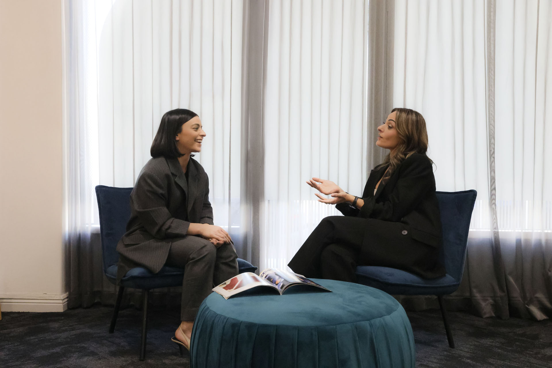 Two women in business attire sitting in the Resolve Divorce Adelaide family law office, engaged in conversation across a teal ottoman with a magazine on top.