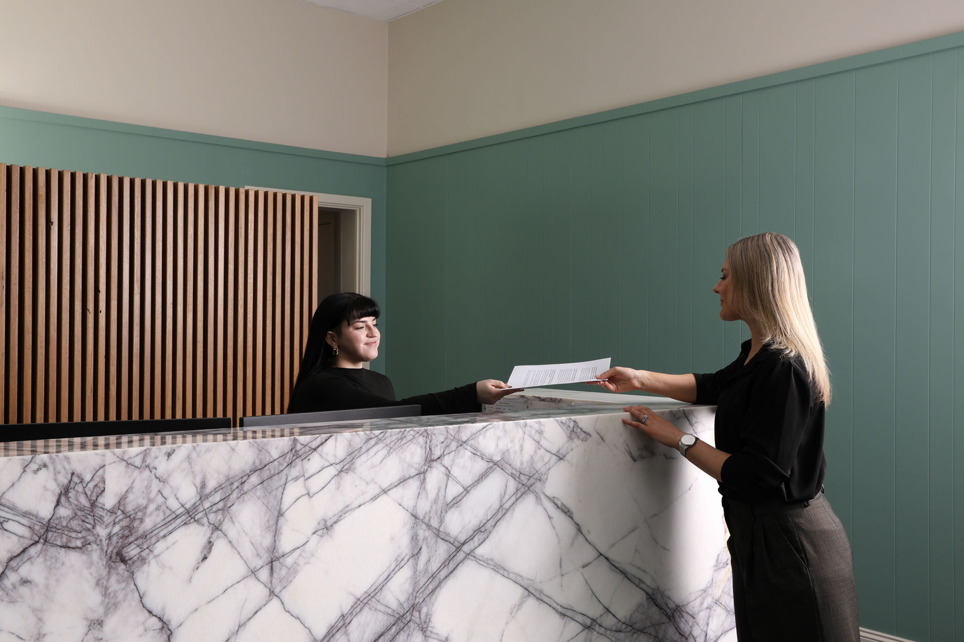 Receptionist at Resolve Divorce, a leading Adelaide family law firm, greeting a client across a marble counter with wooden paneling and a teal wall in the background.