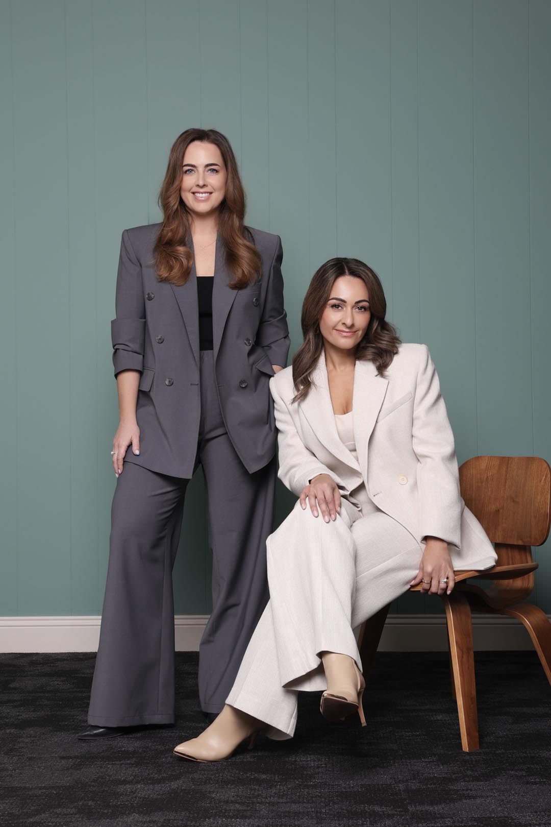 Two women from Resolve Divorce, a leading Adelaide family law firm, one seated and the other sitting confidently in front of a teal wall and marble feature, both dressed in tailored suits and smiling.