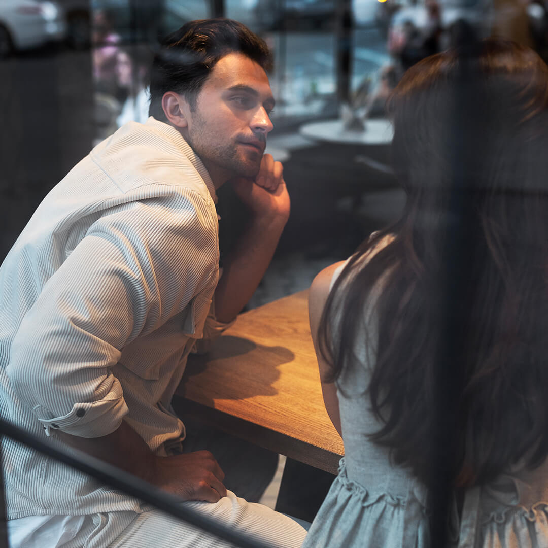 Couple sitting at a café table having a thoughtful conversation, viewed through a window, representing discussions around prenuptial agreements — Resolve Divorce Adelaide family law firm.